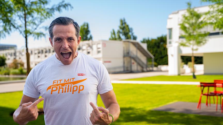 Philipp Jelinek von "Fit mit Philipp" steht draußen und zeigt mit beiden Zeigefingern auf sein weißes T-Shirt mit dem Logo von "Fit mit Philipp". 