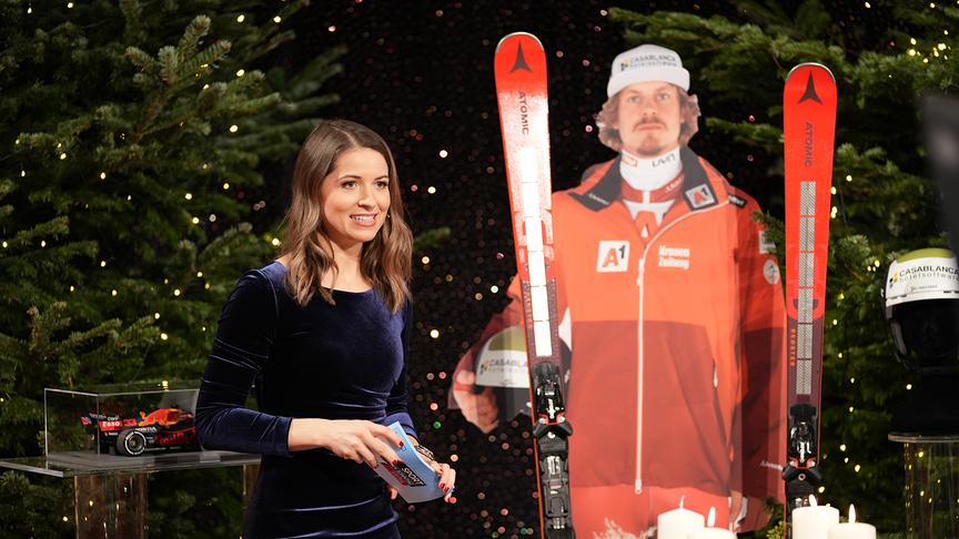 Die Moderatorin Kristina Inhof ist im Bild zu erkennen, außerdem steht ein ausgeschnittener Skifahrer als aufsteller im Bild, auf diesem wurden Skier angelehnt.  Der Hintergrund ist mit Tannenbäumen und kleinen weißen Lichter dekoriert. 
