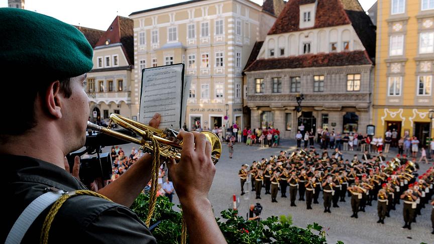 Ein Mann mit Trompete und Liedtext, gekleidet in der grünen Militärmusikuniform und grünem Hut, spielt auf seinem Instrument. Im Hintergrund sind weitere Musikanten der Militärmusik auf einem Dorfplatz zu erkennen.