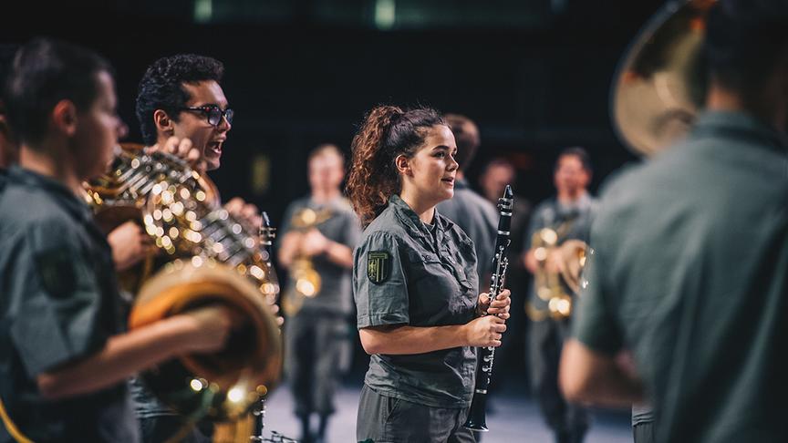 Musikerinnen und Musiker der Militärmusik sind auf diesem Bild abgebildet. Eine junge Frau mit brauen Harren, zusammengebunden zu einem hohen Zopf steht mit einer Klarinette in der Mitte. Weitere Musikanten stehen um Sie herum. Alle Personen tragen eine grüne Uniform. 