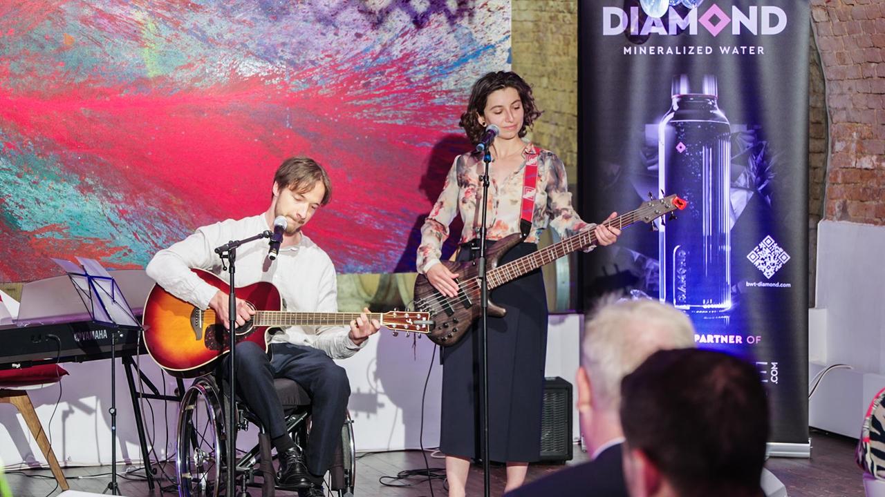 Das Duo "Seidenschwanz" bei seinem Auftritt. Alexander Suppan ist Anfang 30, trägt Bart und spielt in seinem Rollstuhl Gitarre. Sonja Pauli steht neben ihm und spielt auf einer Bassgitarre. Sie ist etwa im gleichen Alter und hat lockige dunkle Haare. Beide haben ein Mikrofon vor sich.
