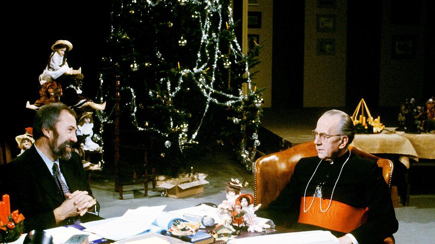 Ernst Wolfram Marboe, ein bärtiger Mann Mitte 40, sitzt an einem Schreibtisch, ihm gegenüber Kardinal Franz König, Ende 70. Das Setting ist weihnachtlich. Der Kardinal trägt eine schwarze Soutane mit roter Paspelierung und ein rotes Zingulum um den Bauch.