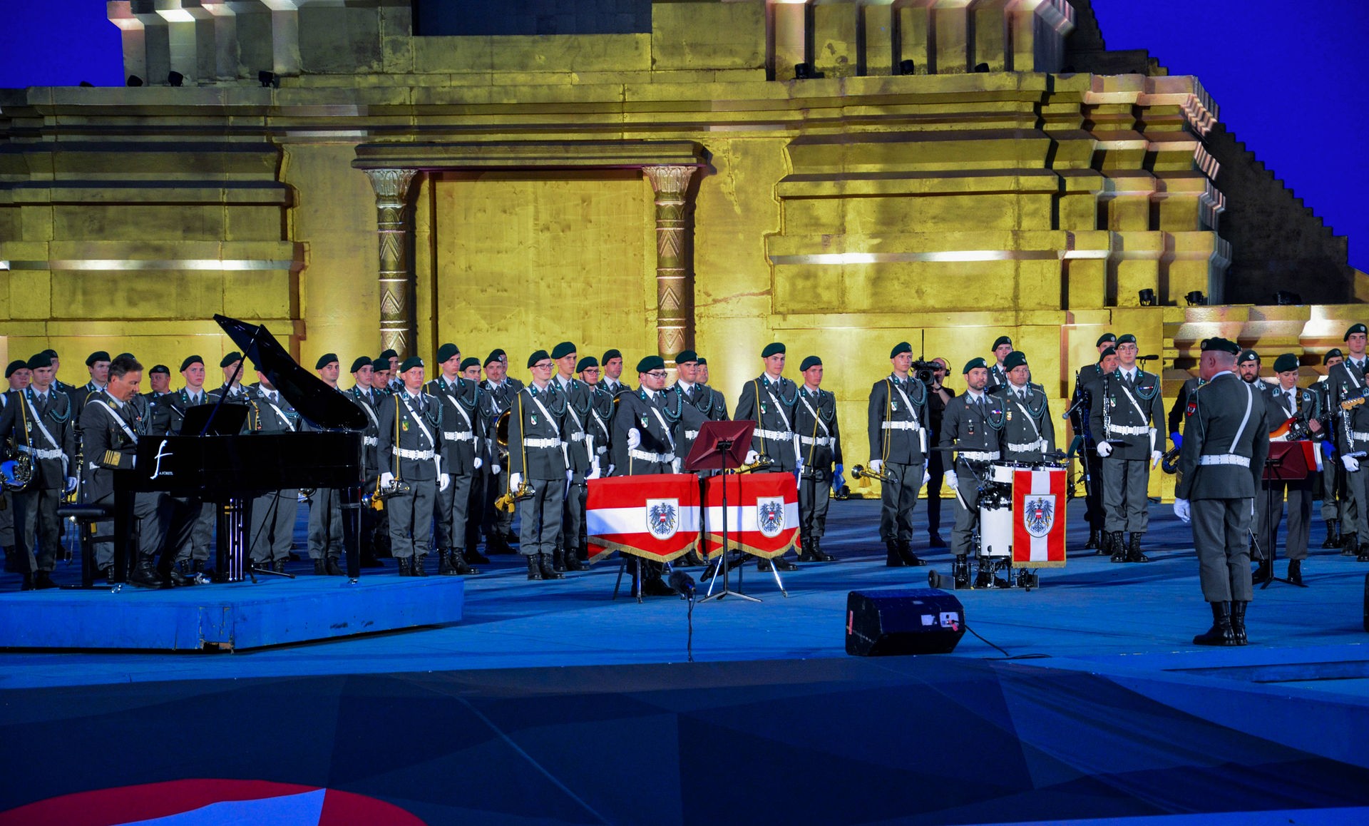 Mehrere Reihen an Musiker*innen in ihrer Gardeuniform stehen auf der Bühne. Im Vordergrund große Trommeln, die mit dem Wappen Österreichs verhängt sind. Links im Bild steht ein schwarzer Klavierflügel.