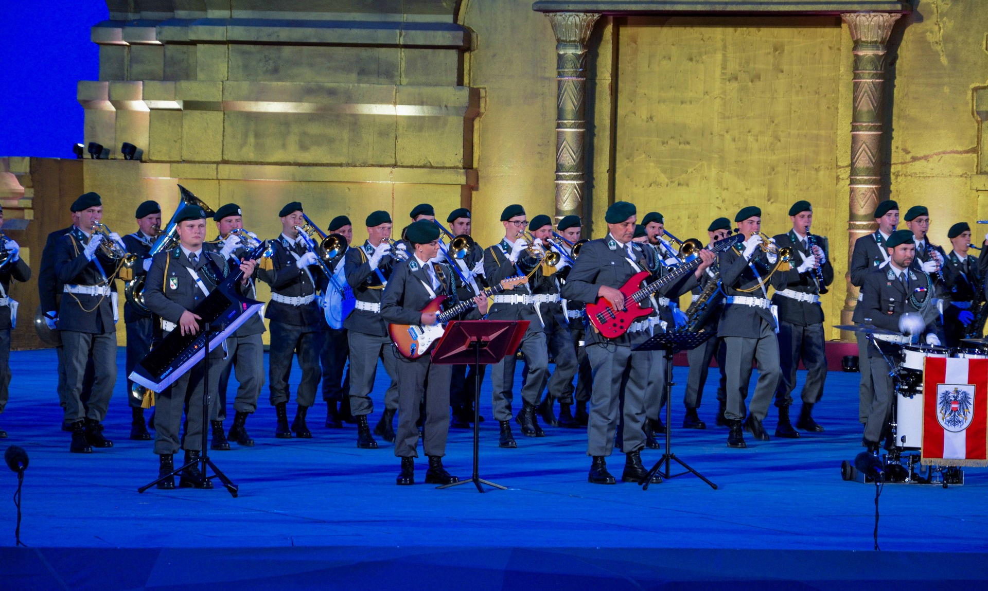 Im Vordergrund sind ein Soldat mit einer schwarzen Keytar, eine Soldatin mit einer orangebraunen E-Gitarre und ein Soldat mit einem roten E-Bass zu sehen. Im Hintergrund stehen mehrere Reihen an Musiker*innen mit Blasinstrumenten.