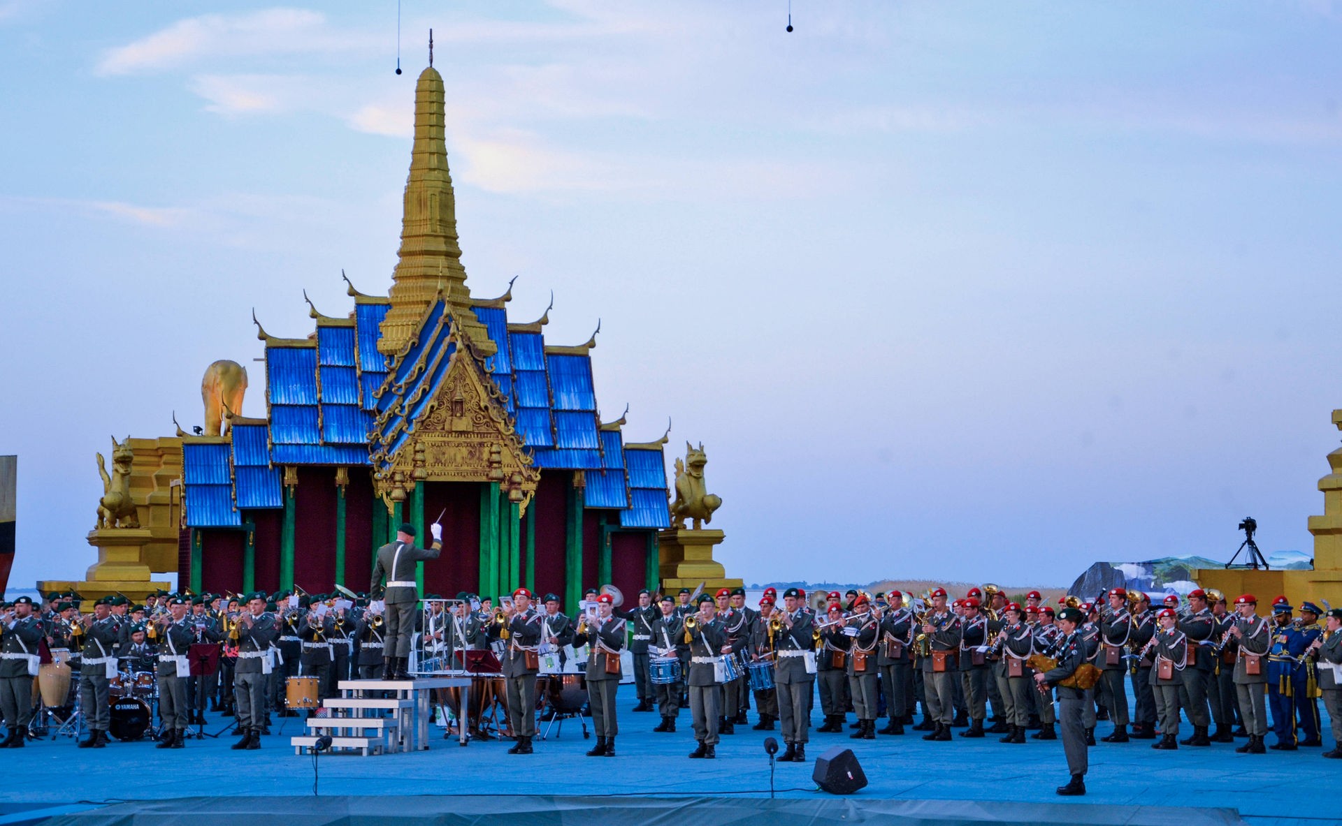 Mehrere Musikkapellen musizieren nebeneinander. Im Vordergrund steht der Dirgent auf einem Podest. Im Hintergrund ist ein Haus mit grünen Säulen und einem blau-goldenen Dach aus dem Bühnenbild von "Der König und Ich" zu sehen.