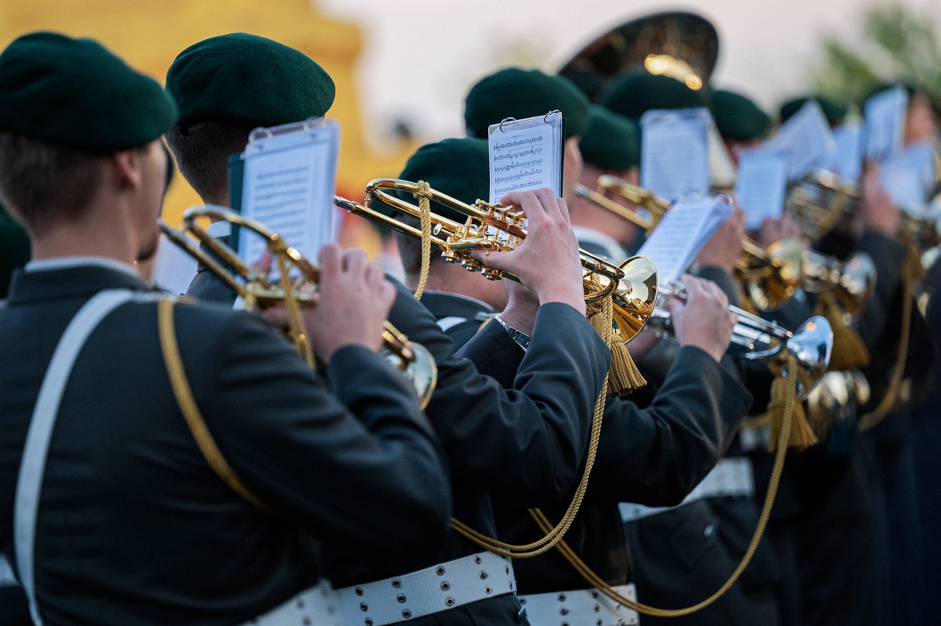 Ein Blick über die Schultern mehrerer Blasmusiker*innen. Sie tragen grüne Kappen, die Instrumente sind goldfarben. Die Noten sind an den Instrumenten angebracht.