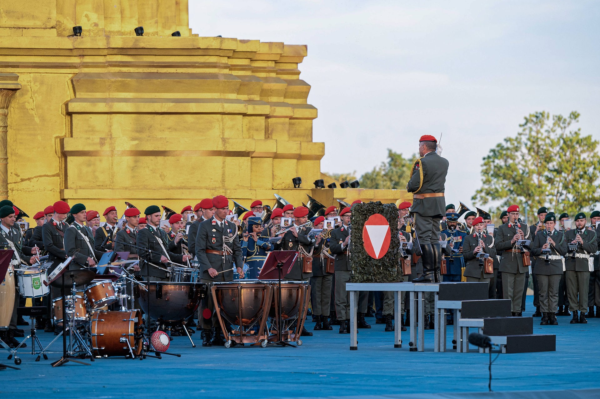 Auf einem Podest steht ein Dirigent mit dem Rücken zum Publikum, vor ihm das Logo des Bundesheeres auf einem Vorhang aus unechten grünen Blättern. Er dirigiert Musikapellen bestehend aus mehreren Dutzend Soldat*innen in Gardeuniform mit roten und grünen Kappen. In der Menge stehen auch eine Soldatin und ein Soldat aus dem Oman, in blauer Gardeuniform mit goldenen Akzenten.