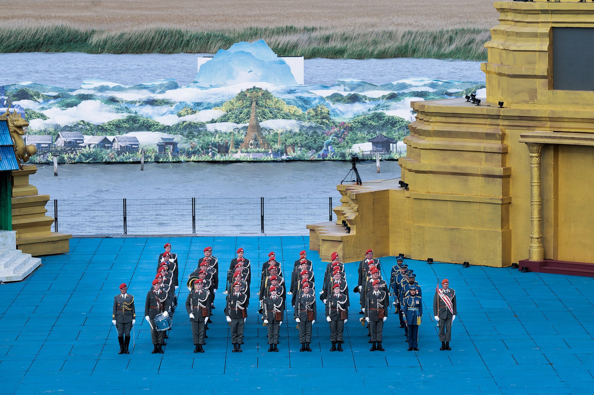 Musiker*innen in grüner Gardeuniform mit roten Kappen und hellen Kordeln stehen in sieben Reihen zu sechs Personen nebeneinander. Rechts von ihnen eine Reihe aus fünf Soldat*innen aus dem Oman. Sie tragen blaue Uniformen mit goldenen Kordeln und Akzenten sowie Hüte anstatt Kappen. Im Hintergrund sind das Bühnenbild von "Der König und Ich" sowie der Neusiedlersee zu sehen.