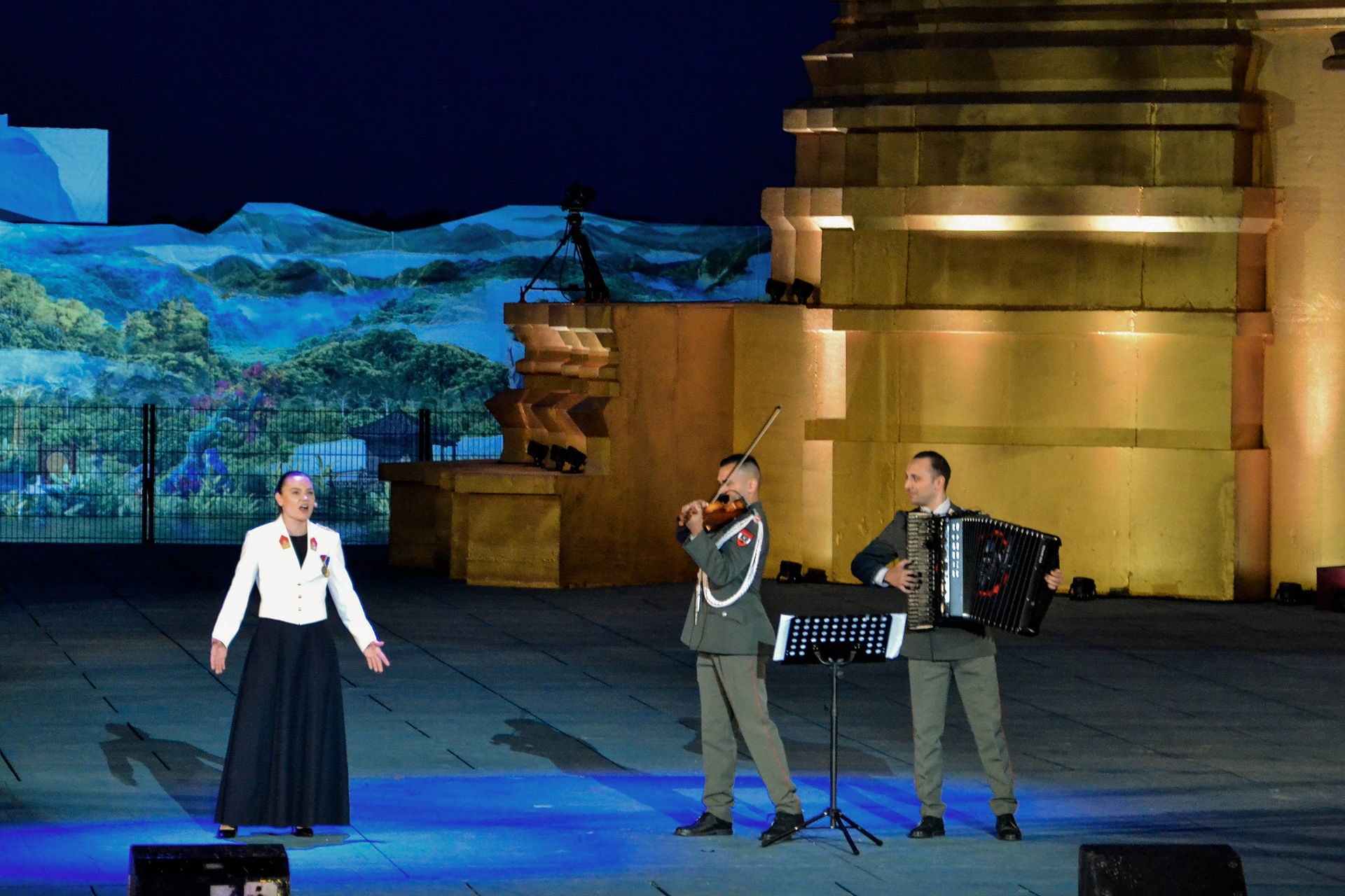 Eine Soldatin in Gala-Uniform - langes, dunkelblaues Kleid und weiße Jacke - steht singend neben zwei Soldaten in Gardeuniform. Einer spielt Violine, der andere Ziehharmonika. 