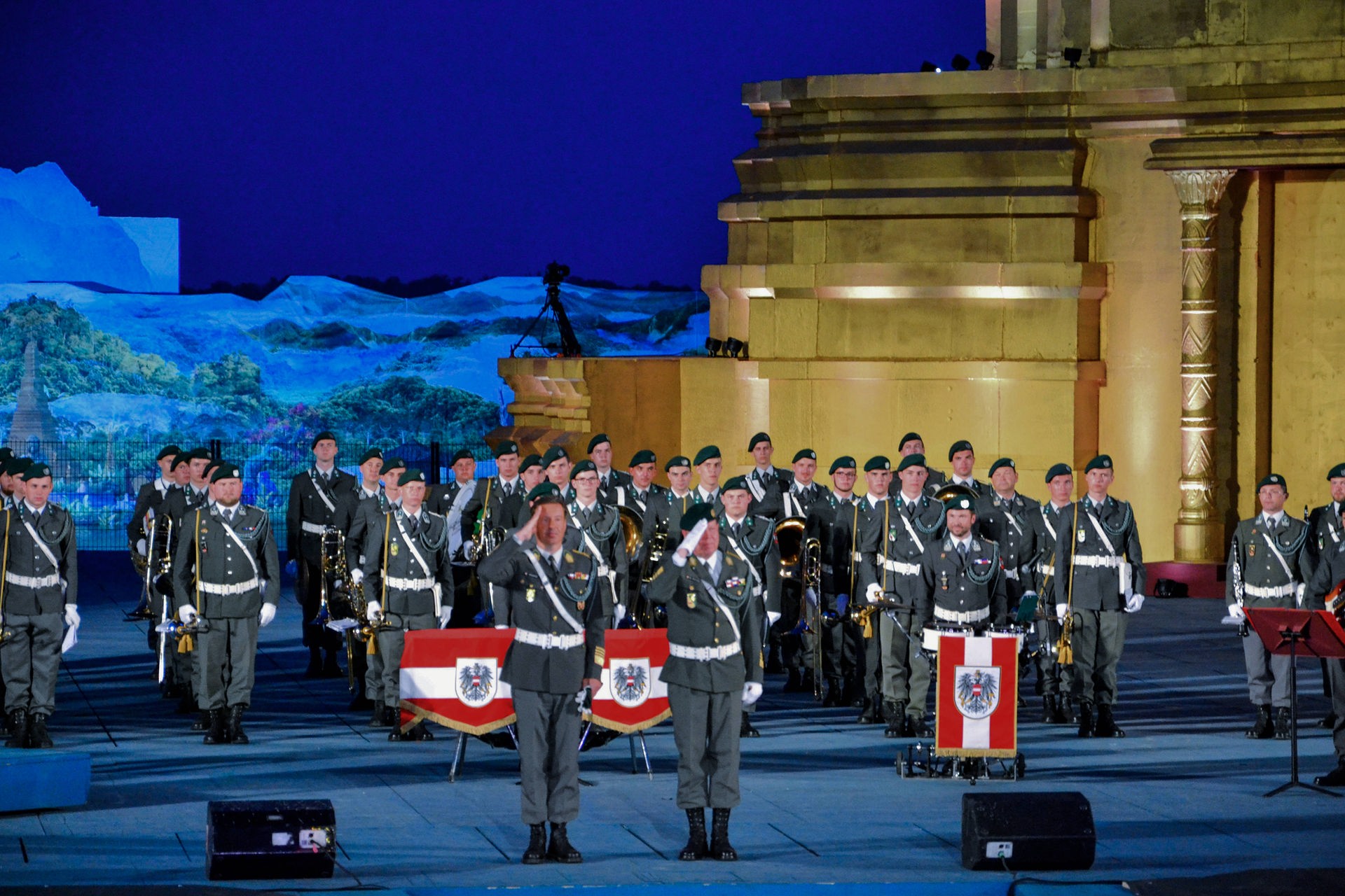 Zwei Soldaten mittig im Bild salutieren. Im Hintergrund eine Musikkapelle in Gardeunifrm mit grünen Kappen und grünen Kordeln. 