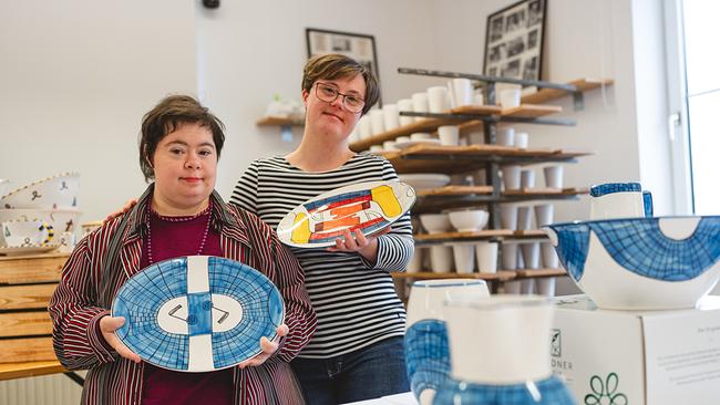 Zwei junge Frauen mit kurzen dunklen Haaren stehen in einer Keramik-Werkstatt und halten bemalte Servierteller in den Händen. Ein Teller ist mit einem Iglu-Eisblockmuster in blau verziert, auf dem anderen sind bunte Eislaufschuhe und Handschuhe zu sehen. Im Hintergrund steht unbemalte Keramik auf Regalen. Beide Frauen haben das Down Syndrom. Sie lächeln in die Kamera.