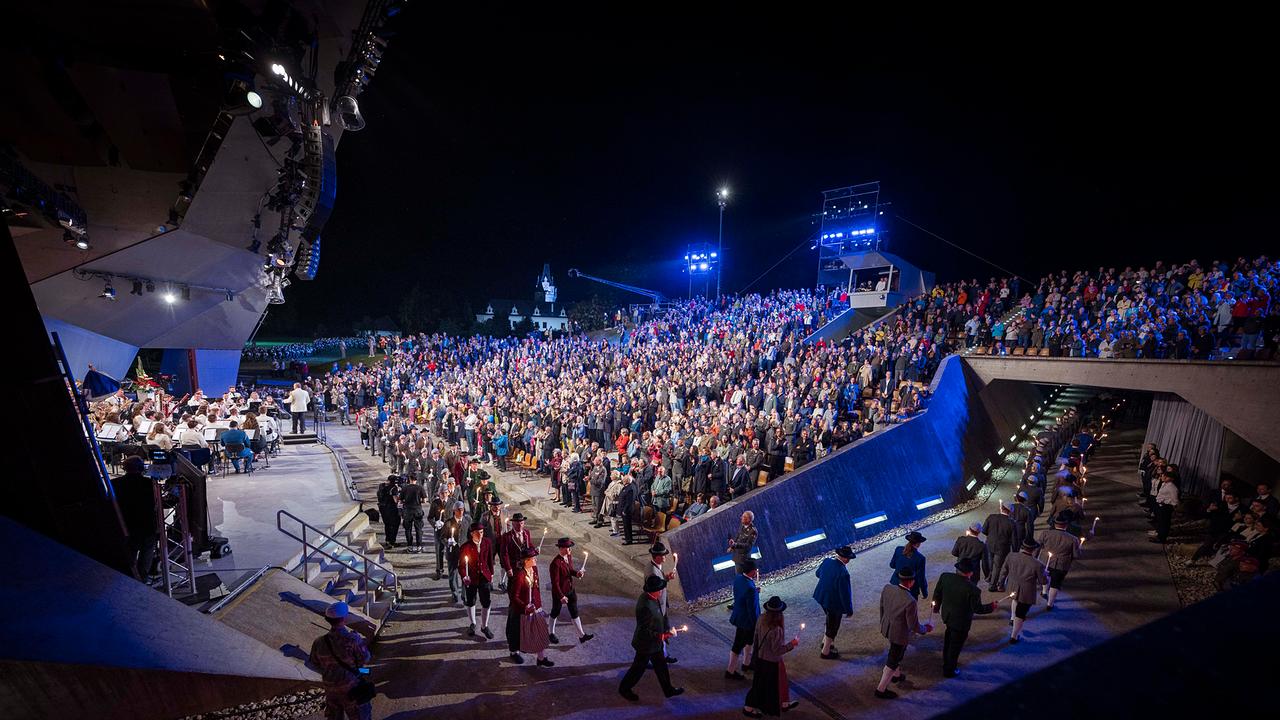 Nachts. Eine große Menschenmenge sitzt in einem Freilufttheater vor einer beleuchteten Bühne. Musiker*innen in Uniform spielen Instrumente, während Personen in traditioneller Kleidung mit Fackeln eine Treppe hinuntergehen.