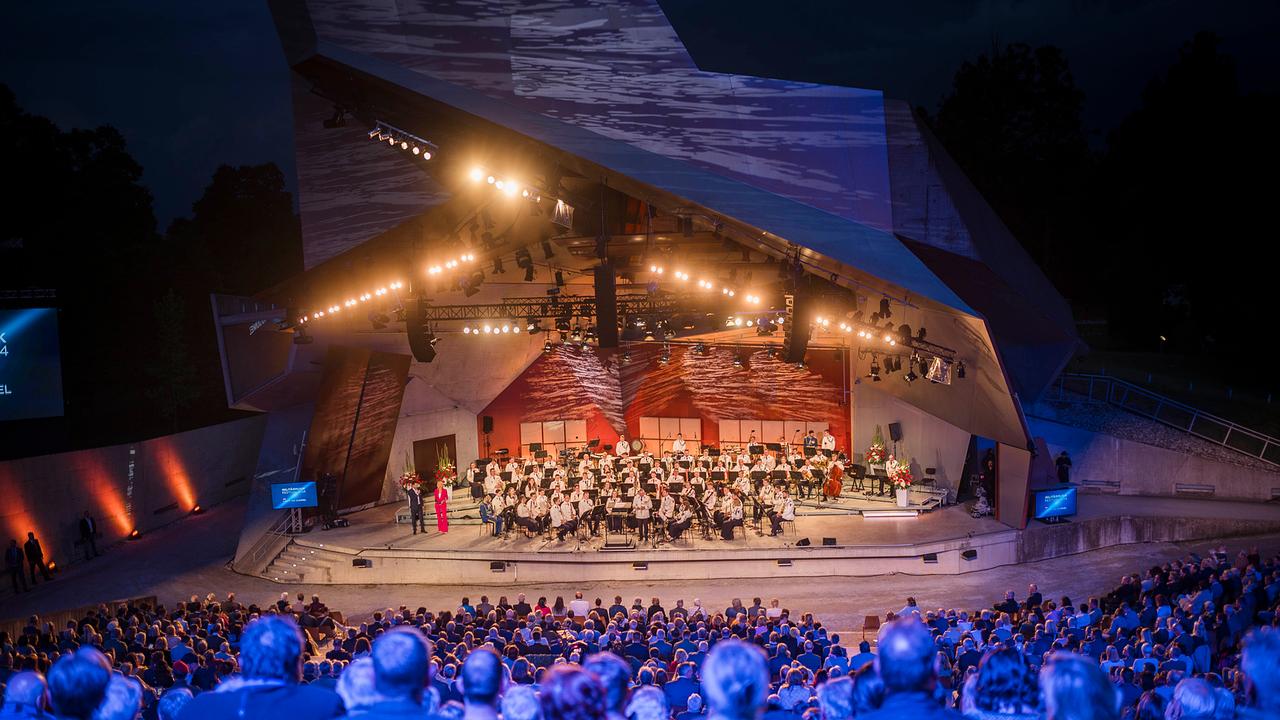 Ein Orchester spielt auf einer modernen, beleuchteten Open-Air-Bühne vor einem großen Publikum. Die Bühne ist von geometrischen Strukturen umgeben, und im Hintergrund ist die Nacht zu sehen.