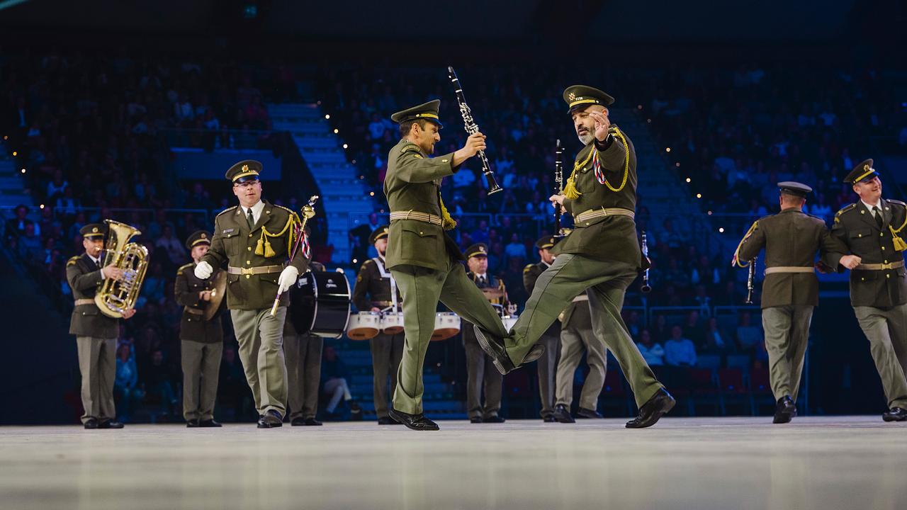 © Mitglieder der tschechischen Militärmusik tanzen mit eingehakten Armen und Beinen fröhlich inmitten ihrer musizierenden Kolleg*innen.