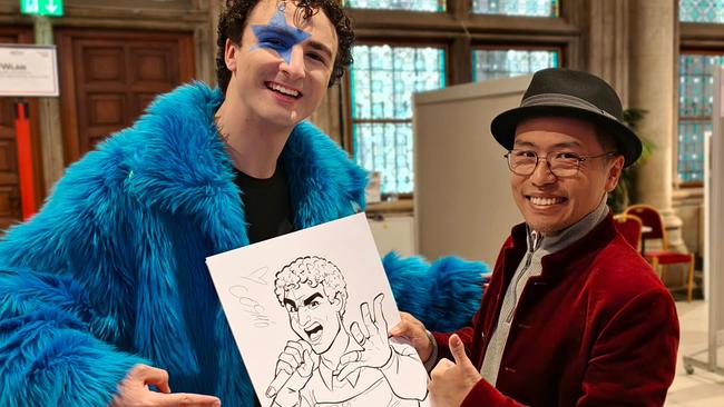 © Xi Ding Cosmo und Xi Ding mit Xi Dings Zeichnung. Cosmo trägt einen blauen Plüschmantel, Xi Ding ein rotes Sakko und einen schwarzen Hut. Die Zeichnung zeigt Cosmo mit Mikrofon in der Hand und ist im Karikatur-Stil gehalten.