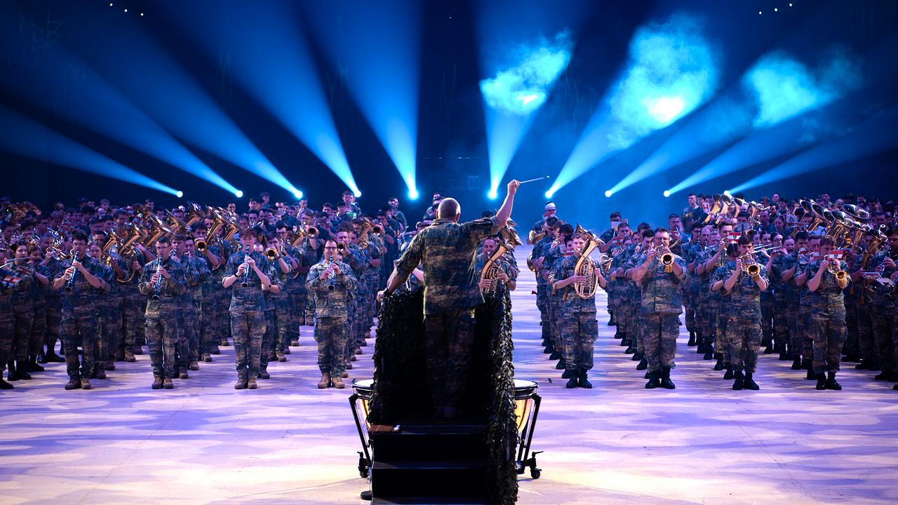 Militärmusikerinnen und Musiker stehen musizierend in mehreren Reihen. Vor ihnen mit dem Rücken zur Kamera der Dirigent, hinter ihnen Nebel- und Lichteffekte. Allen tragen Uniform.