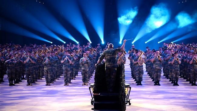 Militärmusikerinnen und Musiker stehen musizierend in mehreren Reihen. Vor ihnen mit dem Rücken zur Kamera der Dirigent, hinter ihnen Nebel- und Lichteffekte. Allen tragen Uniform.