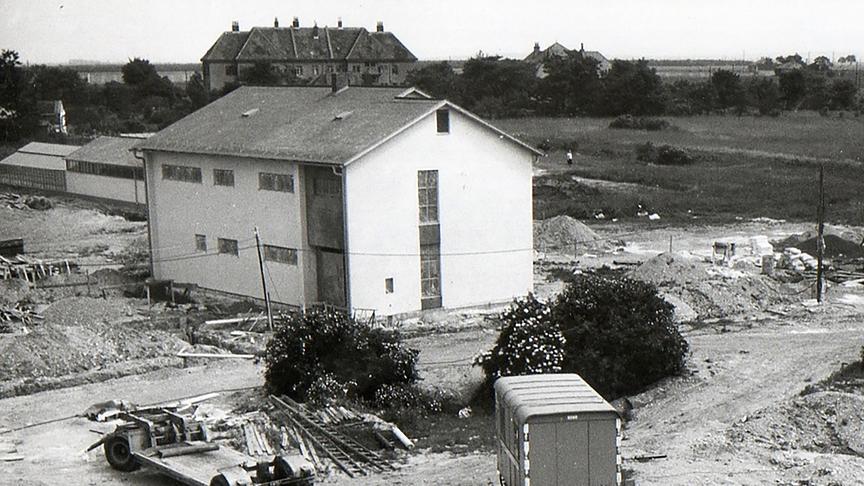 1971 – Aufbau der Sollenau Wohnhäuser. Zu sehen ist eine Baustelle mit einem fertiggestellten Wohnhaus und einem im Bau befindlichen Wohnhaus. Auf der Baustelle befinden sich auch noch Materialien wie Rohre und Leitern und zwei Anhängern, die für die Fertigstellung benötigt werden. Schwarz-weiß-Fotografie