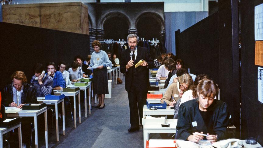 1985 - Ernst Wofram Marboe steht in der Vorbauhalle zwischen den Tischen mit den Telefonierenden und erklärt, wie die Anrufabwicklung in der Spendenzentrale funktioniert.