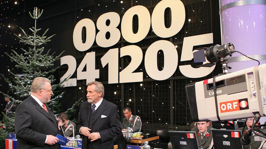 2005 – Elmar Oberhauser und Peter Rapp stehen beide im schwarzen Anzug vor der ORF Kamera. Im Hintergrund sieht man die Telefonnummer von LICHT INS DUNKEL 0800241205 sowie die Gardesoldaten an den Spendentelefonen. Rechts im Bild ist auch eine ORF-Kamera zu sehen.