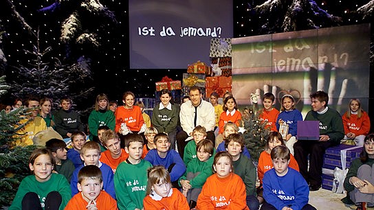 Das Bild zeigt Fernseh-Moderator im Jahr 2000. Armin Assinger sitzt mit Kindern aus diversen Schulen in Österreich im Studio. Die Kinder tragen bunte Sweater mit der Aufschrift "Ist da jemand?"